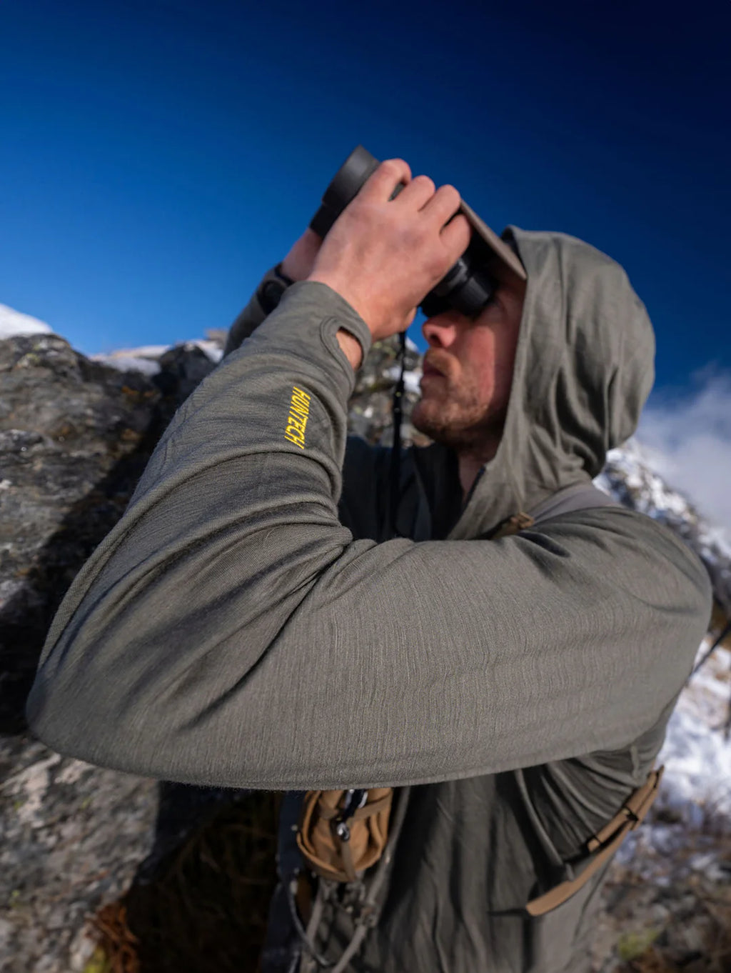 Image of man wearing dark olive green hoodie with the hood up and the Huntech logo in yellow on the left forearm. He's looking through binoculars.