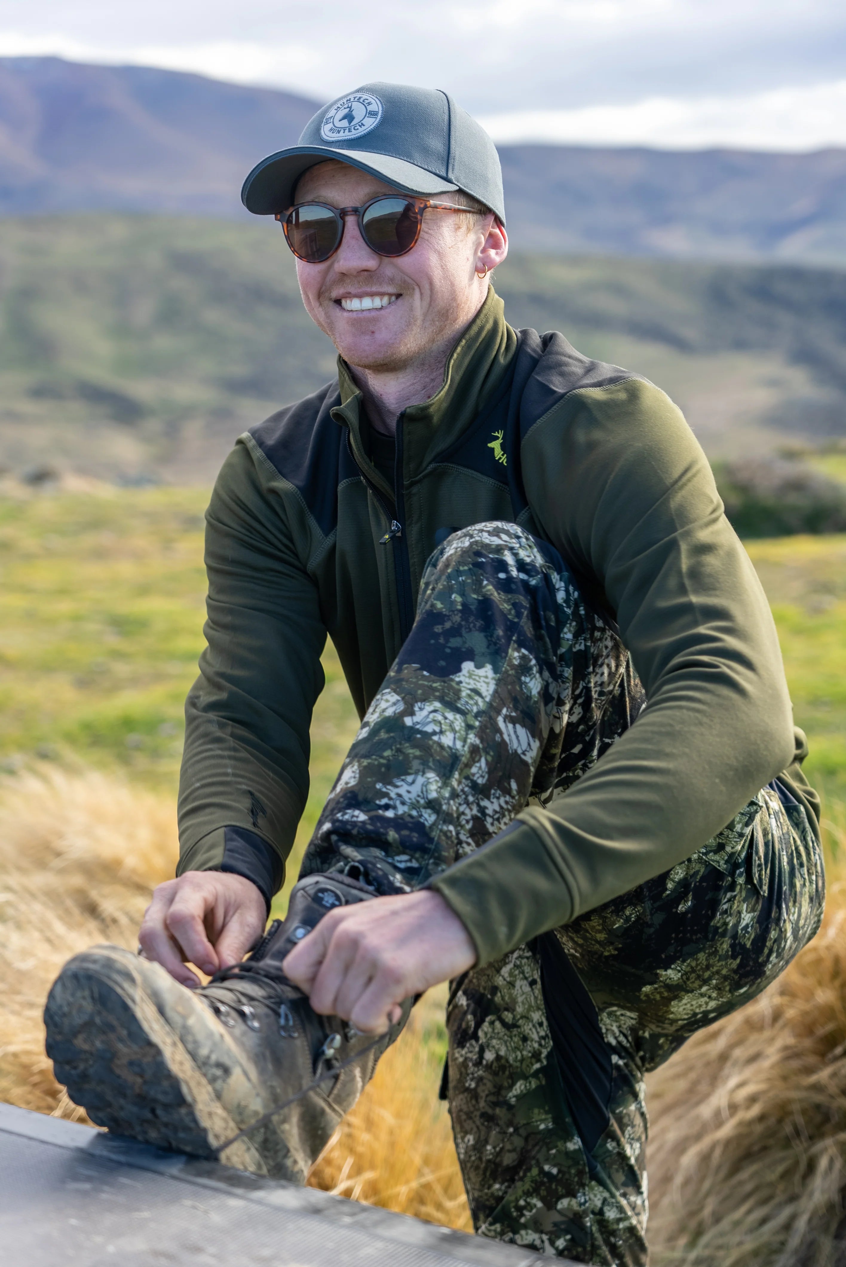 Smiling man tightening the laces on his boots. He is wearing the roar pants in the kakariki camo pattern.