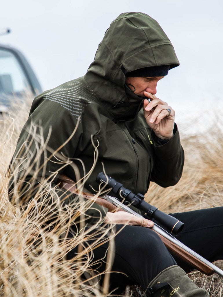 Image of man sitting in tussock carrying a rifle and wearing the jacket in military green.