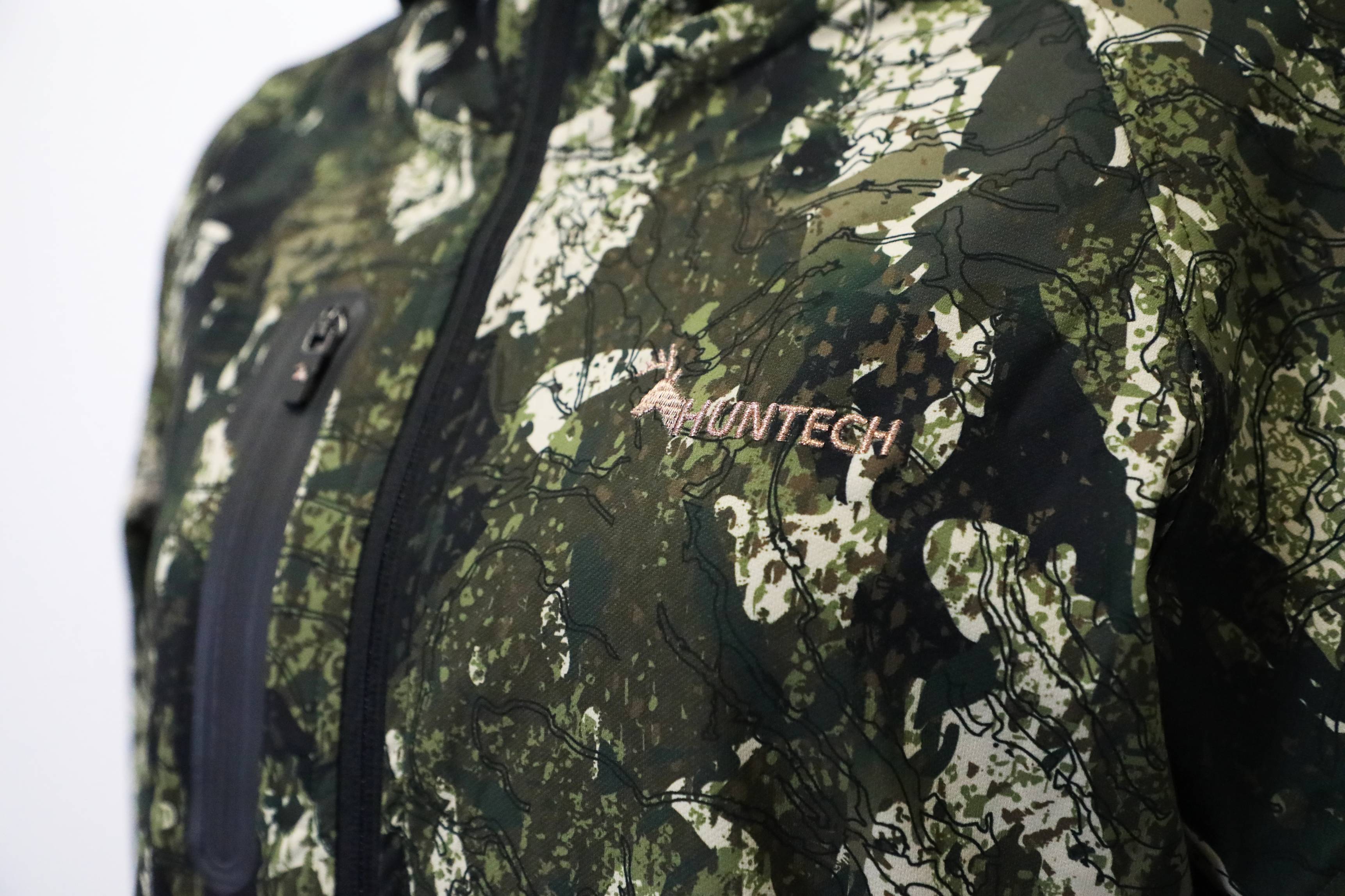 Close up of the Huntech logo in rose gold on the left breast of a zipped up jacket in kakariki camo pattern.
