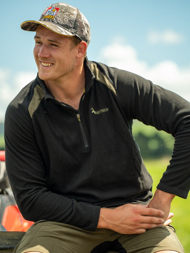 Smiling young man wearing long sleeved top in 180gsm performance fleece with Huntech logo in yellow on the left breast. Zip closed at neck.