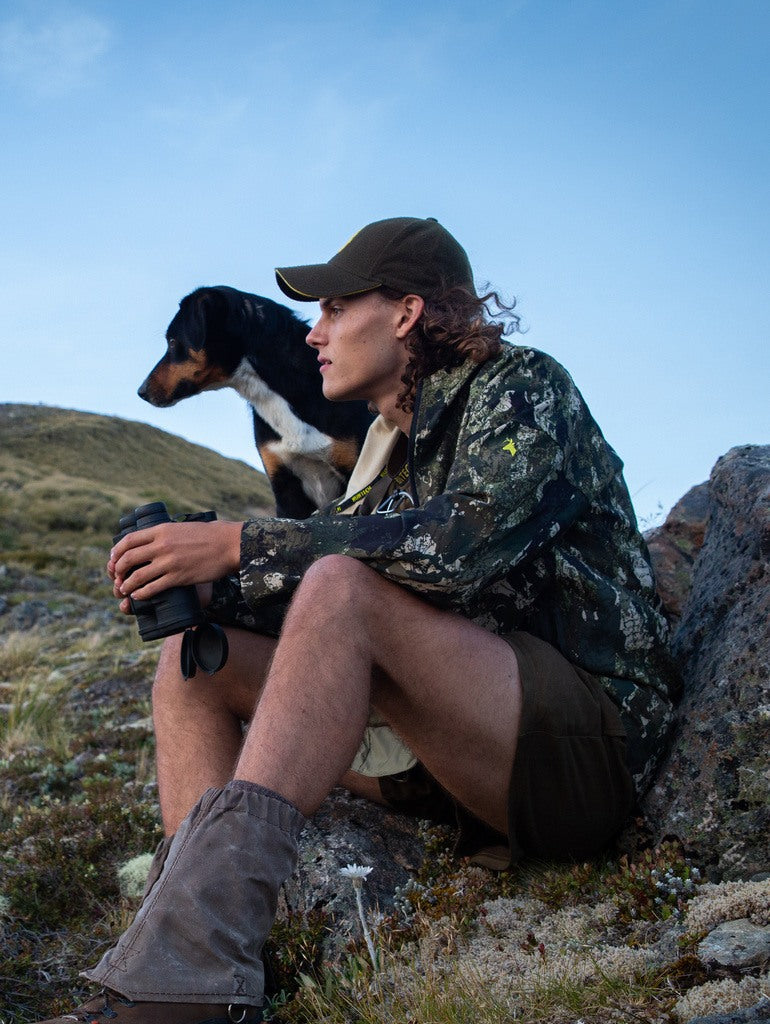 Image of a young man with his dog next to them, both of them intently gazing at something. He is wearing the trail jacket in the kakariki camo pattern with the logo on the left sleeve visible. He is holding binoculars.