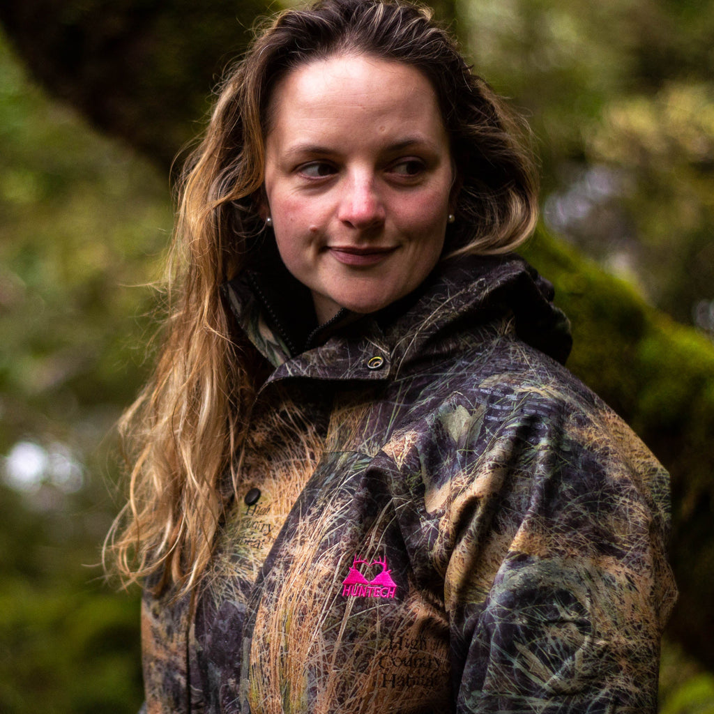 Image of a young woman with shoulder length dark blonde to brunette wearing the tussock jacket in high country camo pattern. She has the hood down and is looking off to the left.