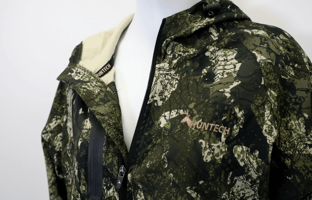 Close up of the Huntech logo in rose gold on the left breast. Zip is partially undone to reveal cream fleece inner. Jacket is shown in kakariki camo pattern.