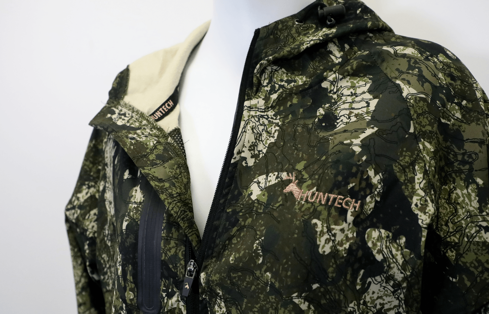 Close up of the Huntech logo in rose gold on the left breast. Zip is partially undone to reveal cream fleece inner. Jacket is shown in kakariki camo pattern.
