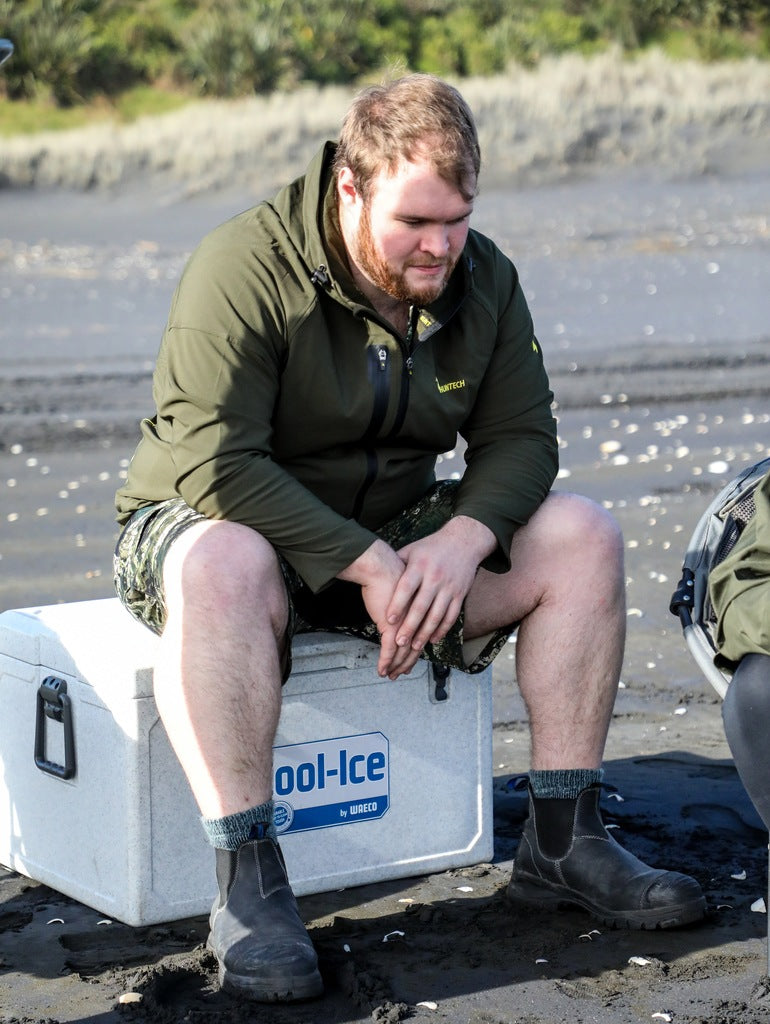 Image of a young man sitting on an Eski and staring pensively at the sandy beach. He is wearing the trail jacket in military green with the Huntech logo on the breast visible.